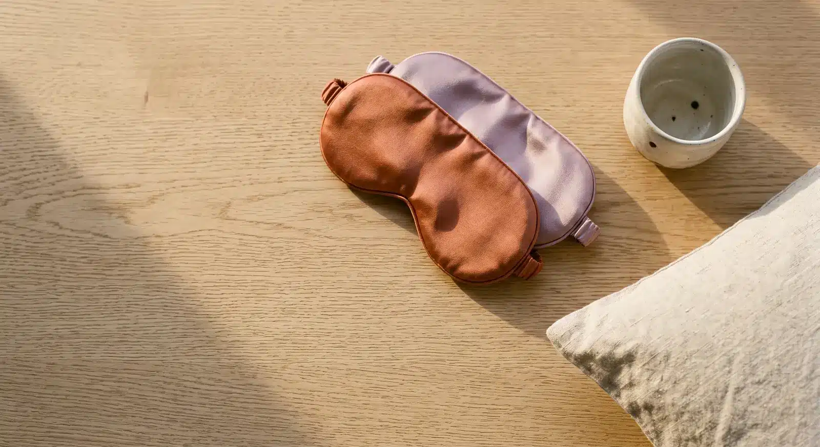 Deux masques de sommeil en satin, terracotta et lavande, à côté d'une tasse et d'un coussin